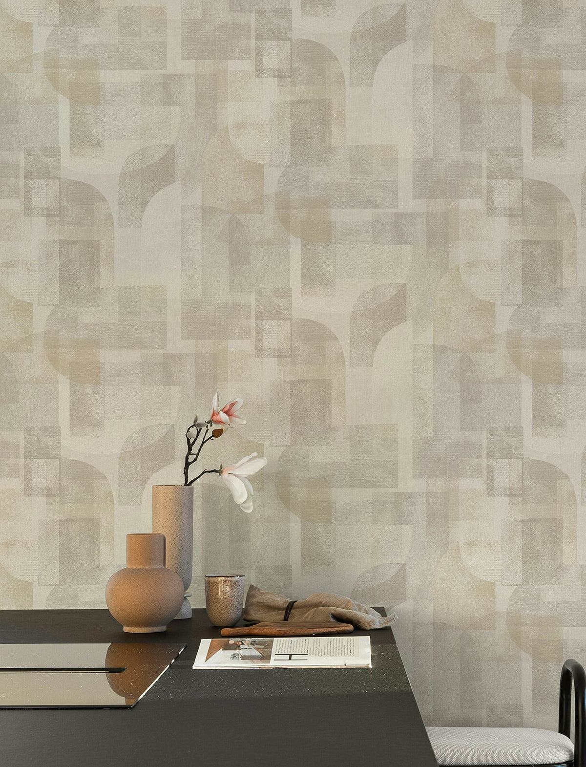 A minimalist table with neutral-toned ceramics, a single branch with white flowers, and papers sits in front of a wall adorned with Chesapeake Cassandra Block Geometric Wallpaper by York Wallcoverings.