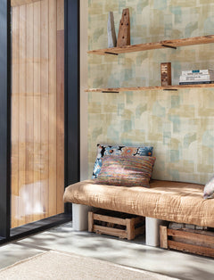 A cozy nook with a tan cushion and colorful pillows on a wooden bench is set against Chesapeake Cassandra Block Geometric Wallpaper by York Wallcoverings, with wooden shelves holding books and decor. Sunlight streams in through a floor-to-ceiling window.