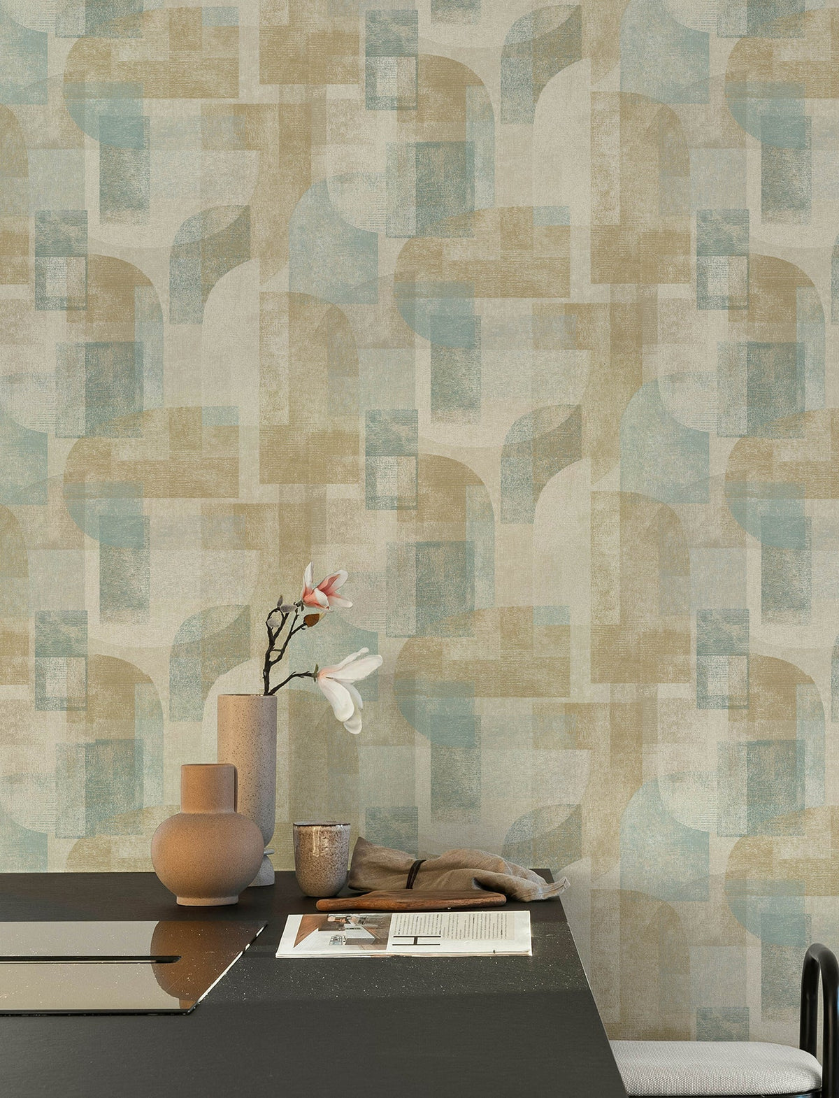 A modern dining area features a black table topped with beige vases, a single flower, a newspaper, and a beige napkin. The walls are adorned with York Wallcoverings’ Chesapeake Cassandra Block Geometric Wallpaper in soft blue and tan abstract geometric patterns.