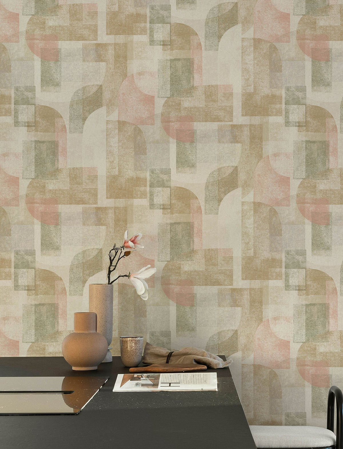 Modern dining table with ceramic vases, a cup, and a flower, set against a wall covered in York Wallcoverings Chesapeake Cassandra Block Geometric Wallpaper in beige, green, and pink. A beige cloth lies on the dark tabletop.