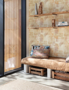 A cozy bench with patterned pillows sits by a large window. Wooden shelves filled with books and decor are mounted on a wall adorned with York Wallcoverings Chesapeake Cassandra Block Geometric Wallpaper. Sunlight streams into the room.