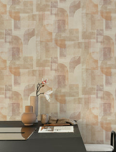 A modern table with neutral-toned vases, a single magnolia branch, and a newspaper sits in front of a wall adorned with York Wallcoverings’ Chesapeake Cassandra Block Geometric Wallpaper in soft shades of beige, peach, and mauve.