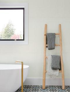 A minimalist bathroom featuring York Wallcoverings’ Chesapeake Sonia Faux Linen Wallpaper, a white tub with a gold faucet, a patterned tile floor, a wooden ladder displaying gray and white towels, and a window showcasing tree views.