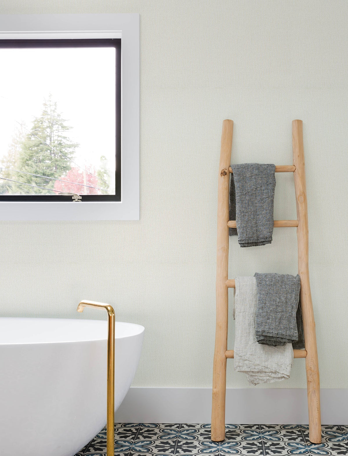 A wooden ladder holding three folded towels stands next to a white bathtub with a gold faucet, near a window and decorative tile floor in a bright bathroom accented with Chesapeake Sonia Faux Linen Wallpaper by York Wallcoverings.