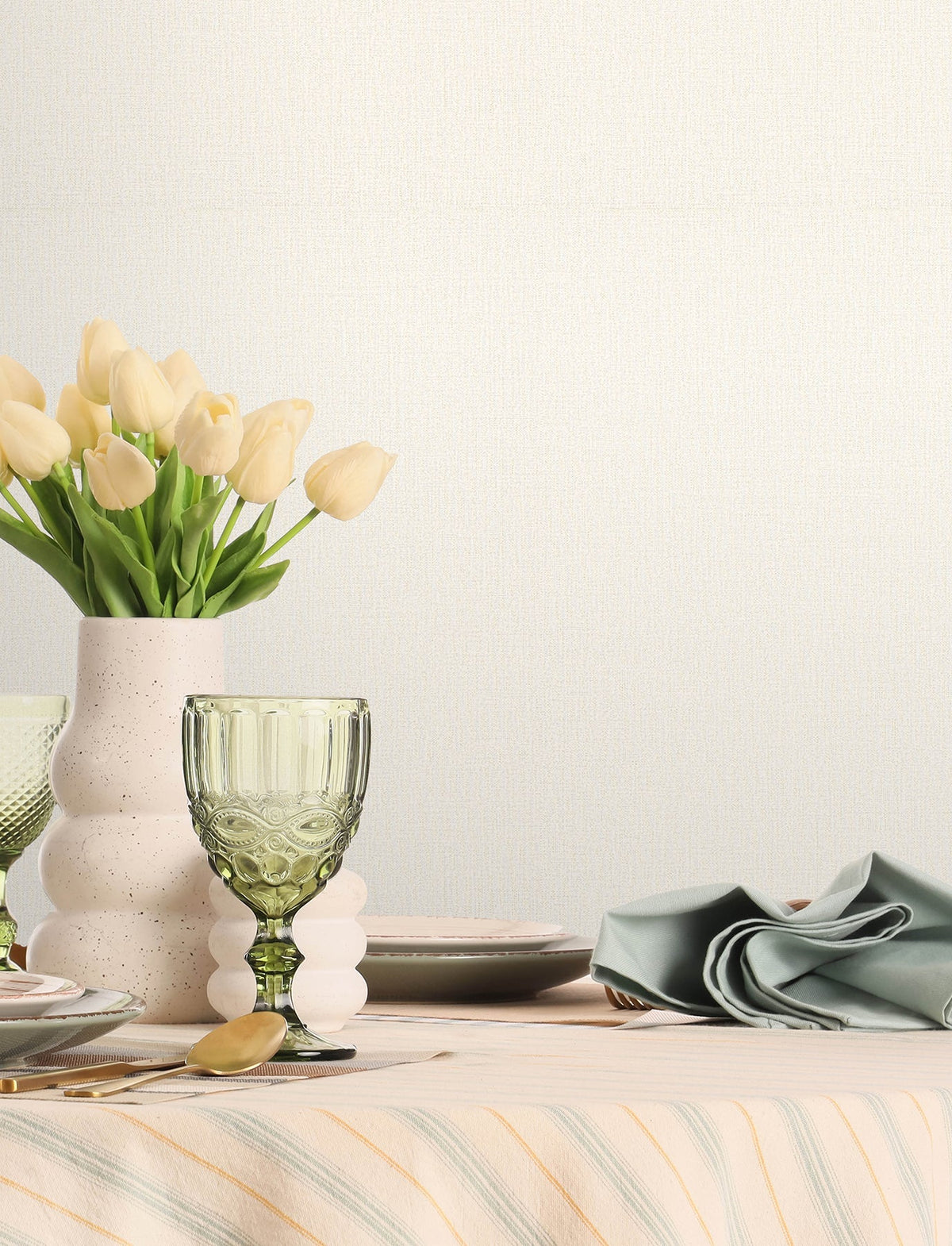 A table set with pastel dishes, a green glass, a folded napkin, and a white vase filled with yellow tulips sits against a backdrop of Chesapeake Sonia Faux Linen Wallpaper by York Wallcoverings.
