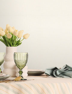 A table set with pastel dishes, a green glass, a folded napkin, and a white vase filled with yellow tulips sits against a backdrop of Chesapeake Sonia Faux Linen Wallpaper by York Wallcoverings.