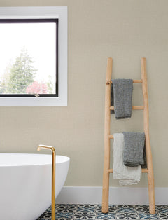A modern bathroom featuring Chesapeake Sonia Faux Linen Wallpaper by York Wallcoverings, a freestanding white tub with a gold faucet, a wooden ladder holding three towels, patterned floor tiles, and a window revealing trees outside.