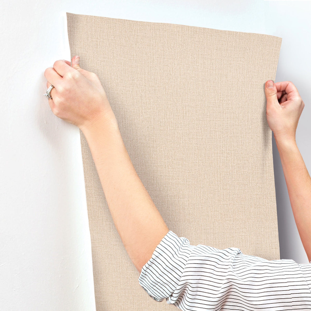 A person wearing a striped shirt is applying Chesapeake Sonia Faux Linen Wallpaper by York Wallcoverings—a beige, textured wallpaper—to a white wall, smoothing it with their hands.