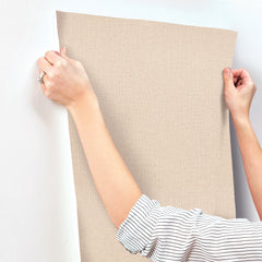 A person wearing a striped shirt is applying Chesapeake Sonia Faux Linen Wallpaper by York Wallcoverings—a beige, textured wallpaper—to a white wall, smoothing it with their hands.