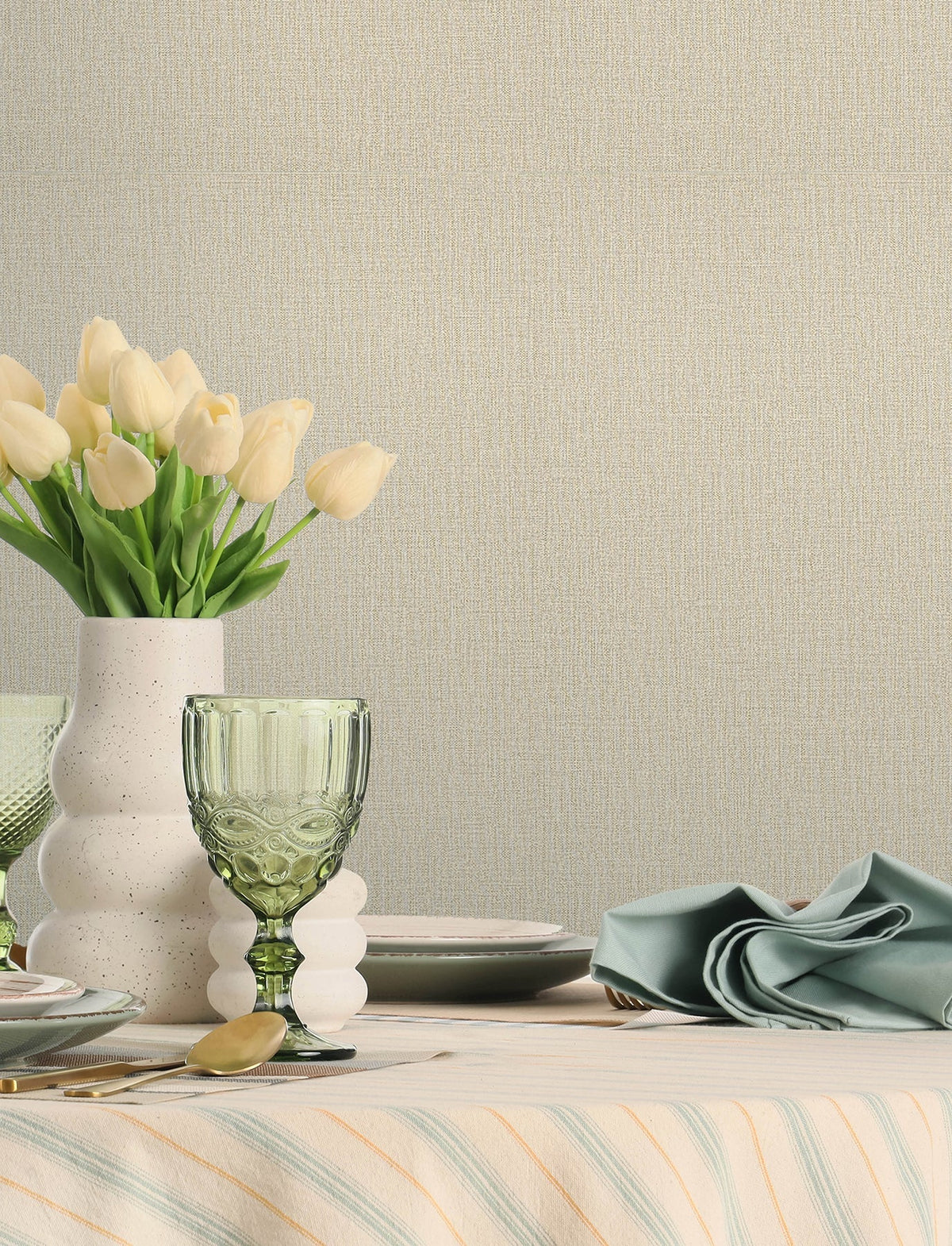 A table set with green glassware, a white speckled vase holding cream tulips, stacked plates, cutlery, and a neatly folded sage napkin on a striped tablecloth is styled against a neutral wall adorned with Chesapeake Sonia Faux Linen Wallpaper by York Wallcoverings.