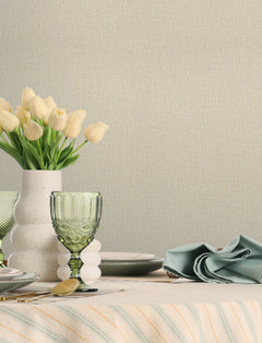 A table set with green glassware, a white speckled vase holding cream tulips, stacked plates, cutlery, and a neatly folded sage napkin on a striped tablecloth is styled against a neutral wall adorned with Chesapeake Sonia Faux Linen Wallpaper by York Wallcoverings.