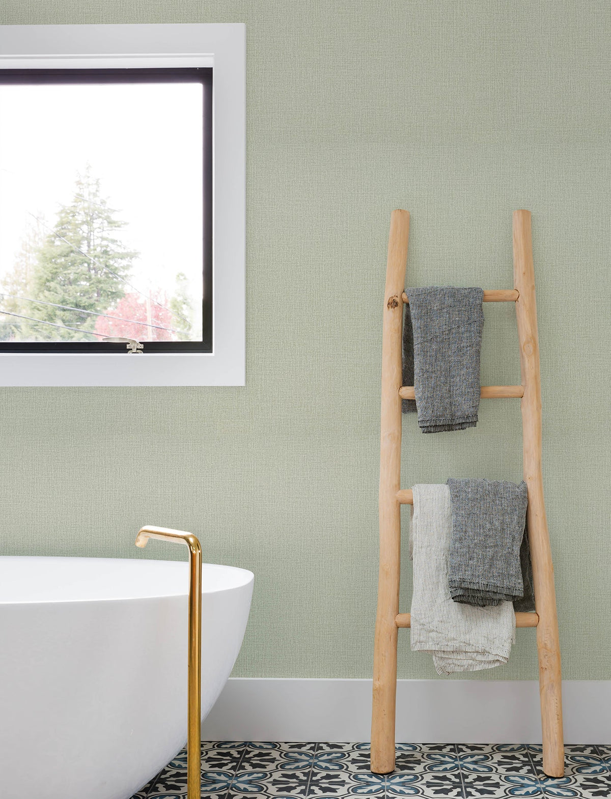 A modern bathroom with a white tub, gold faucet, patterned tile floor, a window, and a wooden ladder holding two towels sits against walls adorned with Chesapeake Sonia Faux Linen Wallpaper by York Wallcoverings.