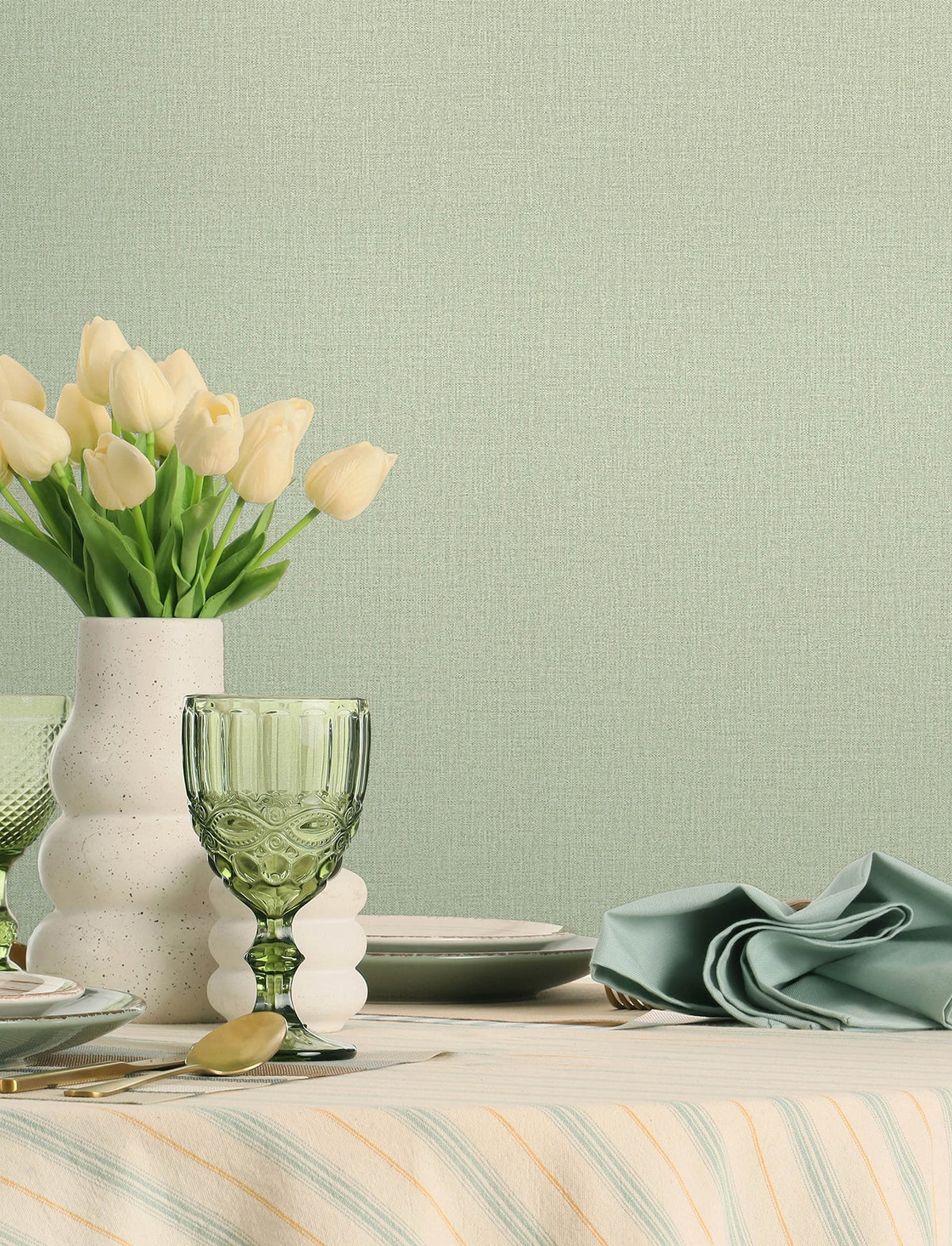A table set with green glassware, a white vase of cream tulips, plates, cutlery, and a folded green napkin sits atop a striped tablecloth, set against walls adorned with Chesapeake Sonia Faux Linen Wallpaper by York Wallcoverings.