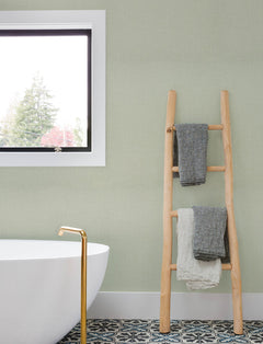 A modern bathroom featuring a white tub, gold faucet, patterned floor tiles, and a wooden ladder holding folded towels sits against walls adorned with Chesapeake Sonia Faux Linen Wallpaper by York Wallcoverings, beside a window that reveals trees outside.