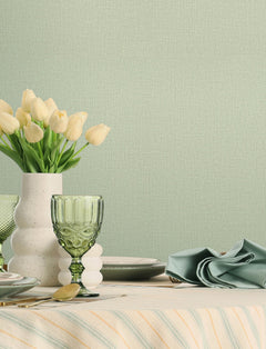 A table set with green glassware, a white vase of pale yellow tulips, plates, cutlery, and a folded green napkin is arranged against a pale green textured wall covered in York Wallcoverings’ Chesapeake Sonia Faux Linen Wallpaper.