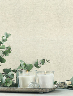 Two lit white candles in glass holders sit on a silver tray with eucalyptus branches and a clear crystal, set against a light beige Chesapeake Tansy Distressed Wallpaper by York Wallcoverings.