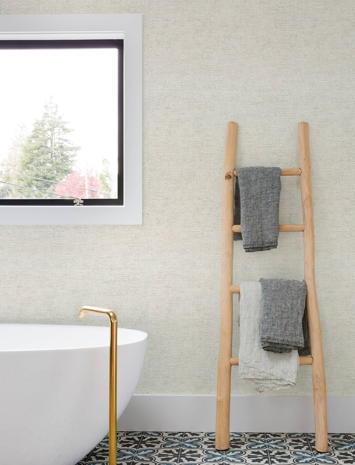 A modern bathroom featuring a white bathtub, gold faucet, patterned tile floor, a window, and a wooden ladder with two folded towels is set against walls adorned in Chesapeake Tansy Distressed Wallpaper by York Wallcoverings.