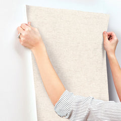 A person in a striped shirt holds and positions a roll of Chesapeake Tansy Distressed Wallpaper by York Wallcoverings against a white wall, preparing to hang it.