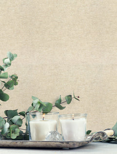 Two lit white candles in glass holders rest on a silver tray with green eucalyptus leaves, set against a neutral beige Chesapeake Tansy Distressed Wallpaper by York Wallcoverings.