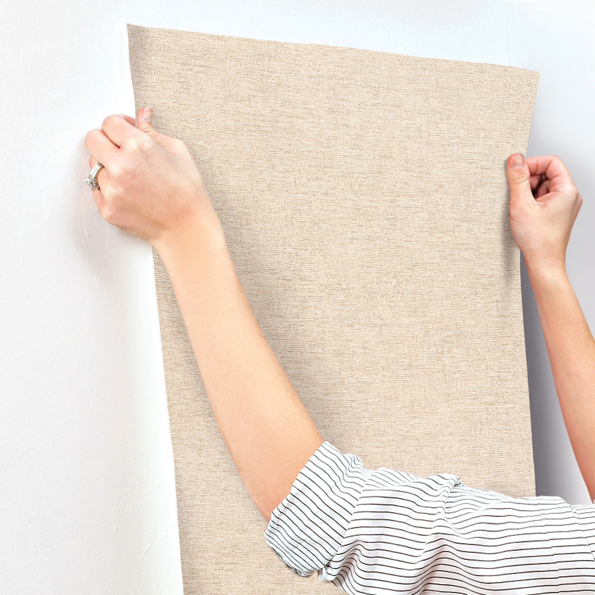 A person in a striped shirt smooths Chesapeake Tansy Distressed Wallpaper by York Wallcoverings against a white wall with both hands.