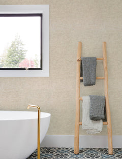 Modern bathroom with a white tub, gold faucet, and a wooden ladder holding three folded towels against a wall covered in Chesapeake Tansy Distressed Wallpaper by York Wallcoverings. Patterned tile floor and a window showing trees outside.