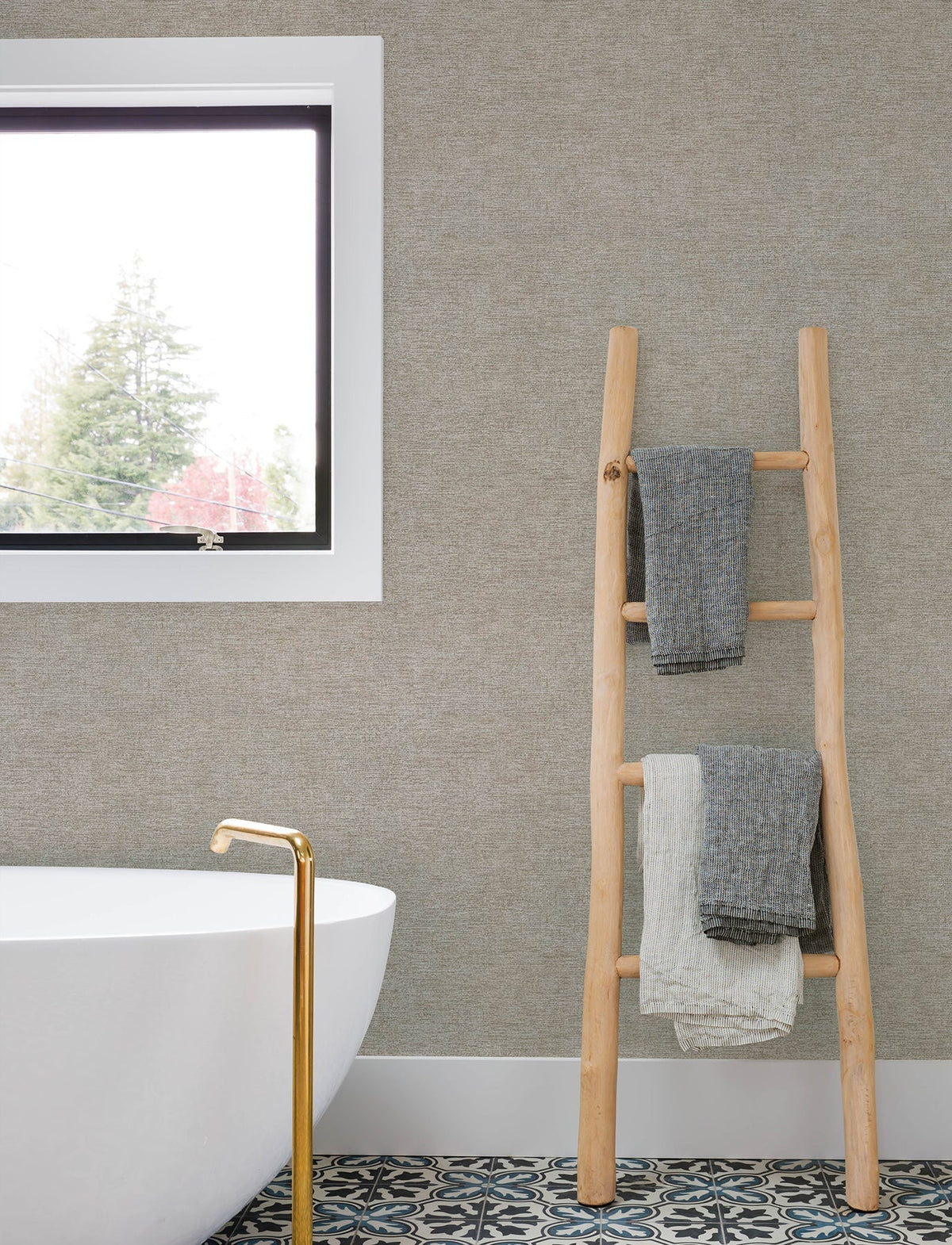 Minimalist bathroom featuring a freestanding white tub, gold faucet, patterned floor tiles, and a wooden ladder displaying two textured towels. The wall above the tub is adorned with Chesapeake Tansy Distressed Wallpaper by York Wallcoverings, with a window offering a tree view.