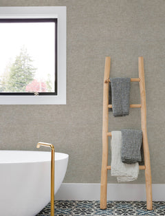 Minimalist bathroom featuring a freestanding white tub, gold faucet, patterned floor tiles, and a wooden ladder displaying two textured towels. The wall above the tub is adorned with Chesapeake Tansy Distressed Wallpaper by York Wallcoverings, with a window offering a tree view.