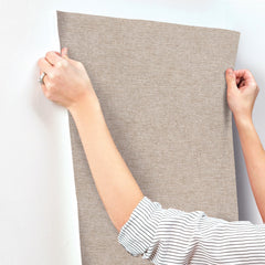 Wearing a striped shirt, a person presses the Chesapeake Tansy Distressed Wallpaper by York Wallcoverings against a white wall, smoothing it with both hands.