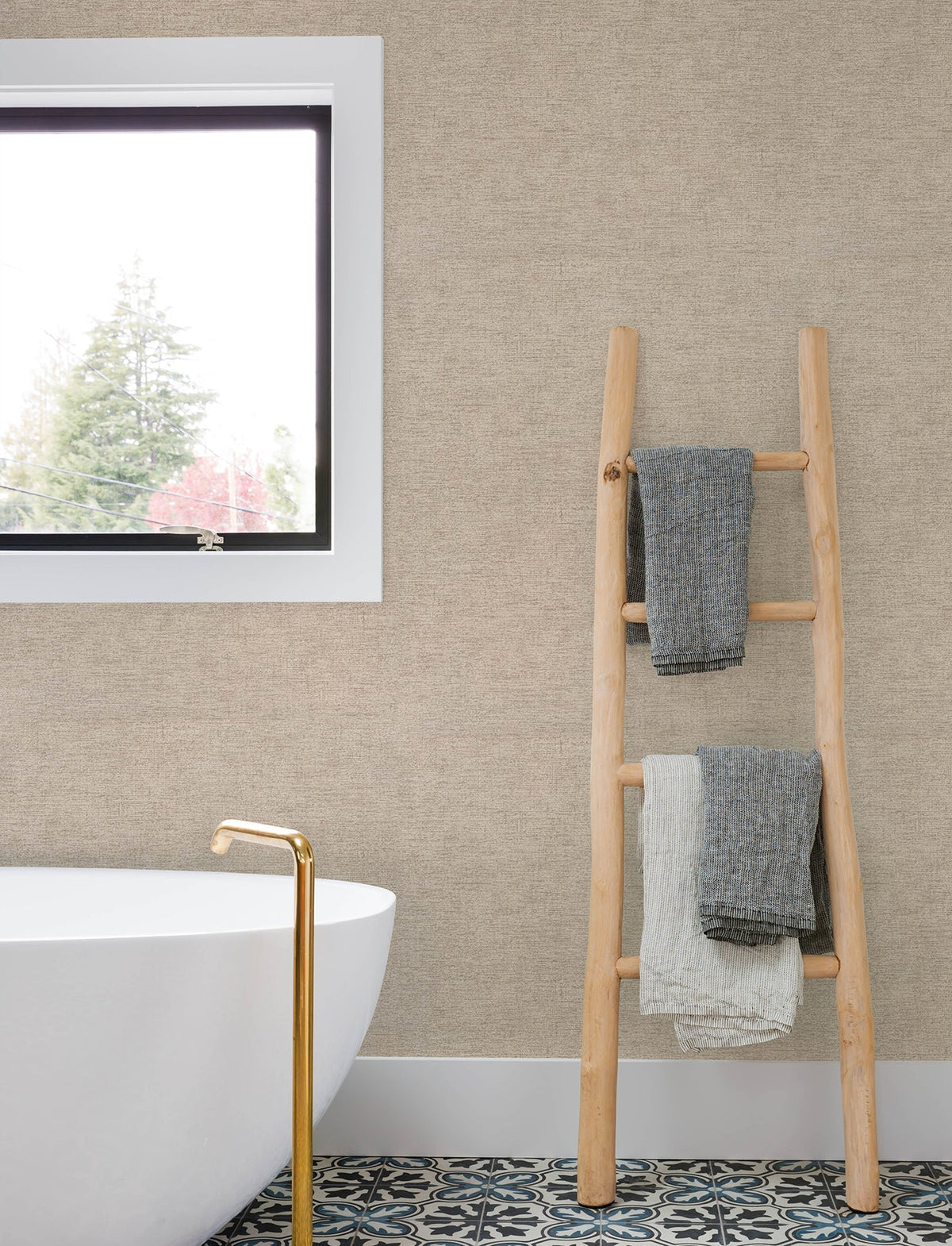 A modern bathroom featuring a white tub, gold faucet, patterned floor tiles, a window, and a wooden ladder holding three folded towels is set against walls adorned with Chesapeake Tansy Distressed Wallpaper by York Wallcoverings.