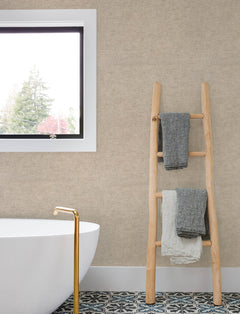 A modern bathroom featuring a white tub, gold faucet, patterned floor tiles, a window, and a wooden ladder holding three folded towels is set against walls adorned with Chesapeake Tansy Distressed Wallpaper by York Wallcoverings.