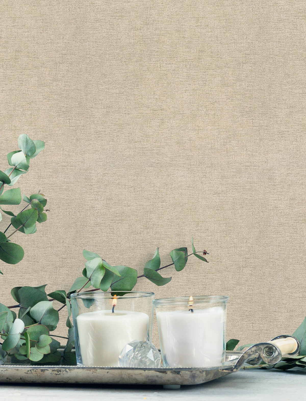 Two white candles in glass holders rest on a silver tray, surrounded by eucalyptus branches, set against York Wallcoverings’ Chesapeake Tansy Distressed Wallpaper.