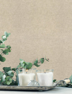 Two white candles in glass holders burn on a decorative silver tray, surrounded by eucalyptus branches, set against Chesapeake Tansy Distressed Wallpaper by York Wallcoverings.