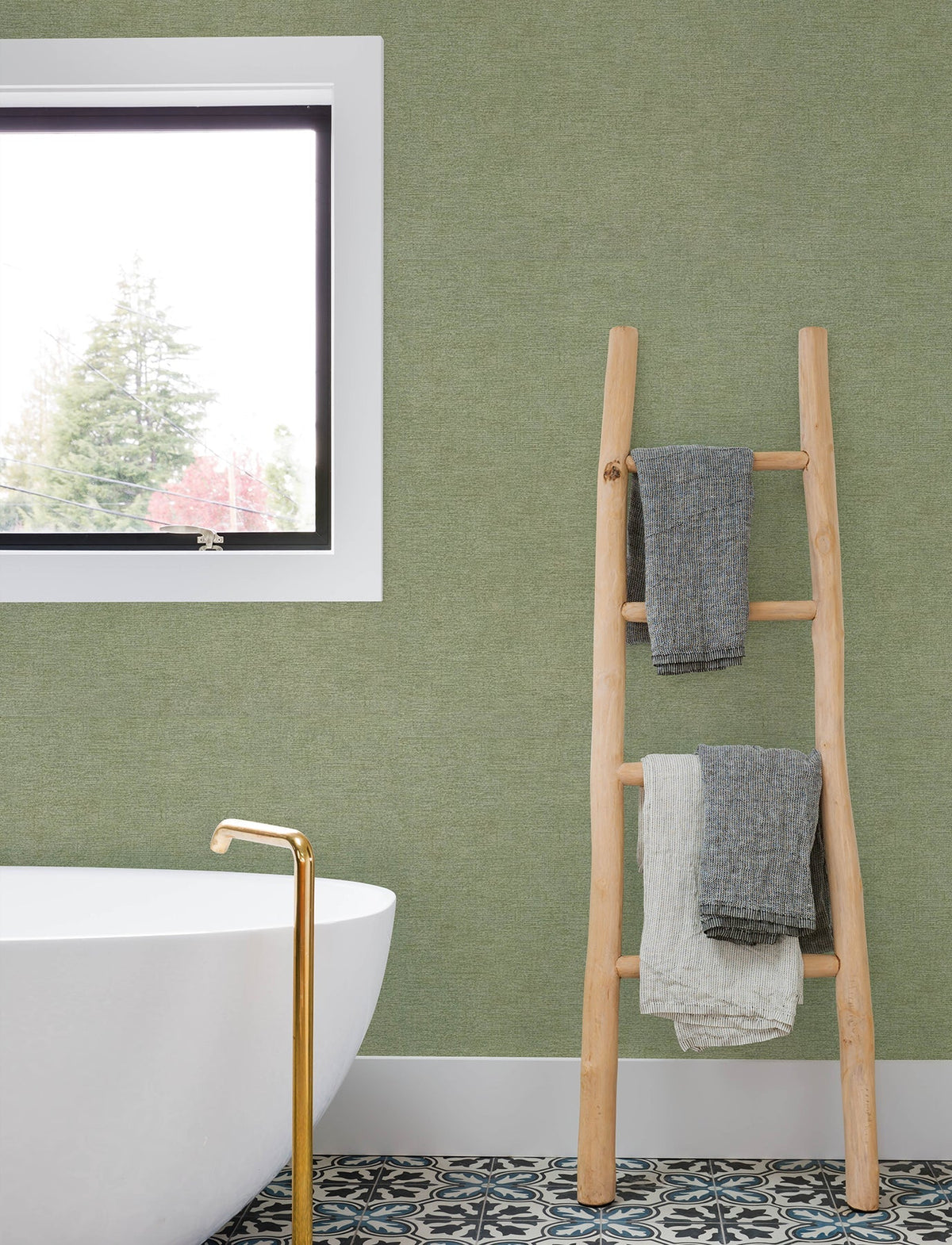 A modern bathroom featuring a freestanding white tub, gold faucet, patterned tile floor, and a window-side wooden ladder holding three folded towels, with the green wall adorned in Chesapeake Tansy Distressed Wallpaper by York Wallcoverings.