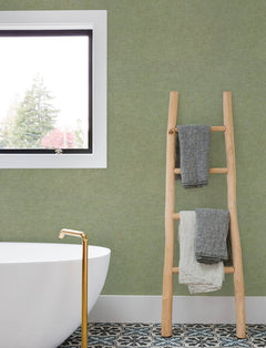 A modern bathroom featuring a freestanding white tub, gold faucet, patterned tile floor, and a window-side wooden ladder holding three folded towels, with the green wall adorned in Chesapeake Tansy Distressed Wallpaper by York Wallcoverings.