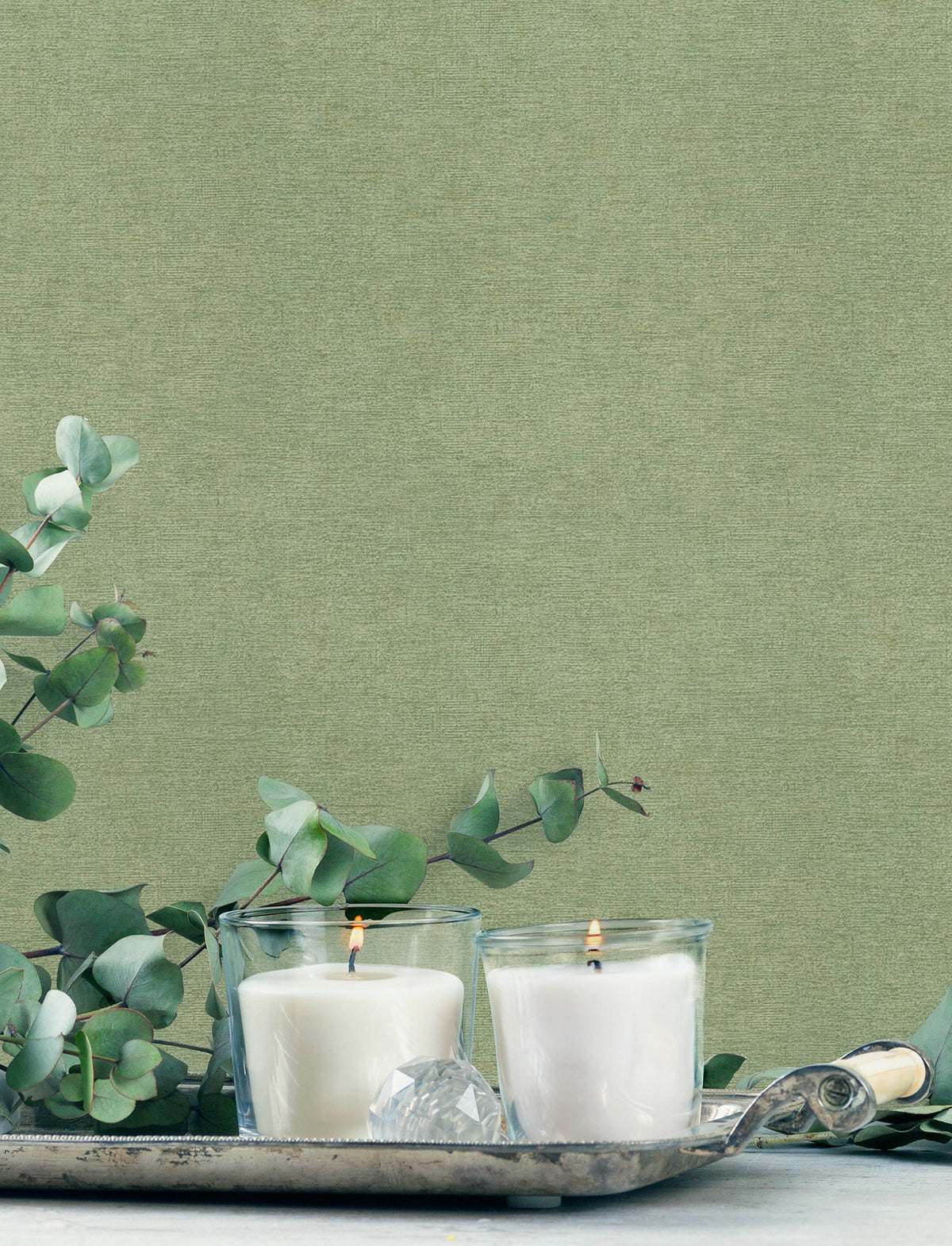 Two white candles in glass holders rest on a silver tray with eucalyptus branches and a faceted clear crystal, set against Chesapeake Tansy Distressed Wallpaper from York Wallcoverings.