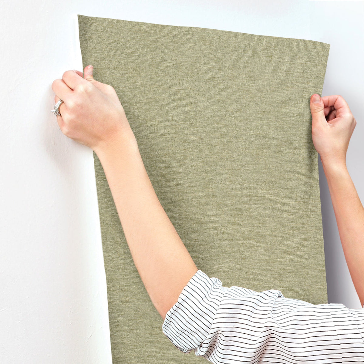A person in a striped shirt is applying Chesapeake Tansy Distressed Wallpaper by York Wallcoverings, smoothing the textured, light-green wallpaper onto a white wall with both hands.