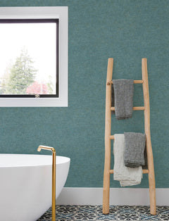 A modern bathroom featuring a white tub, gold faucet, patterned tile floor, wooden ladder with towels, a teal Chesapeake Tansy Distressed Wallpaper by York Wallcoverings on the wall, and a window revealing trees outside.