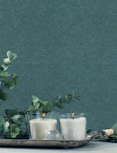 Two white candles in glass holders are lit on a silver tray, surrounded by eucalyptus branches, with York Wallcoverings Chesapeake Tansy Distressed Wallpaper as the blue textured background.