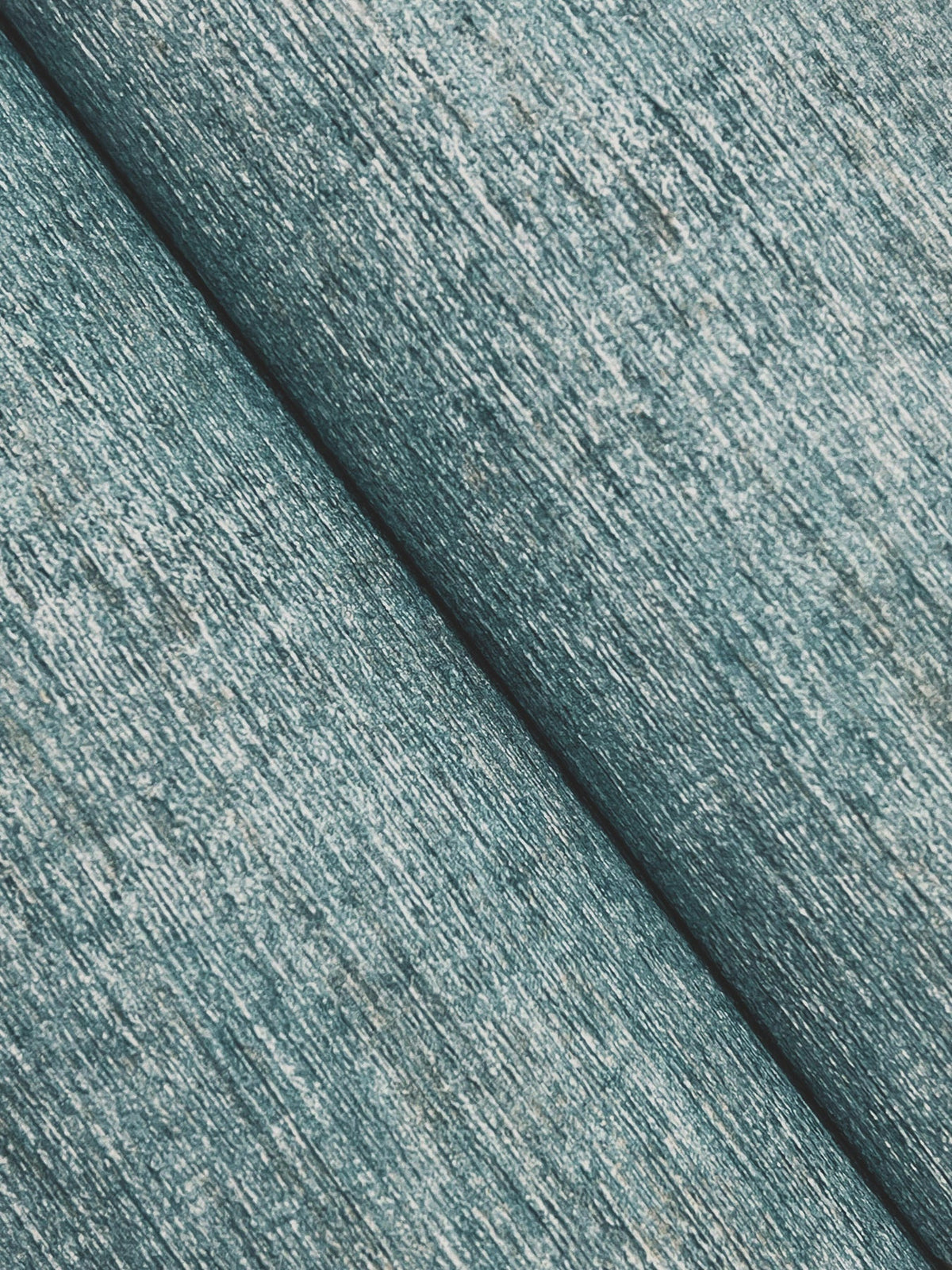 Close-up of Chesapeake Tansy Distressed Wallpaper by York Wallcoverings shows a textured, teal surface with a diagonal seam dividing two panels, highlighting visible fibers and a rustic, distressed finish.