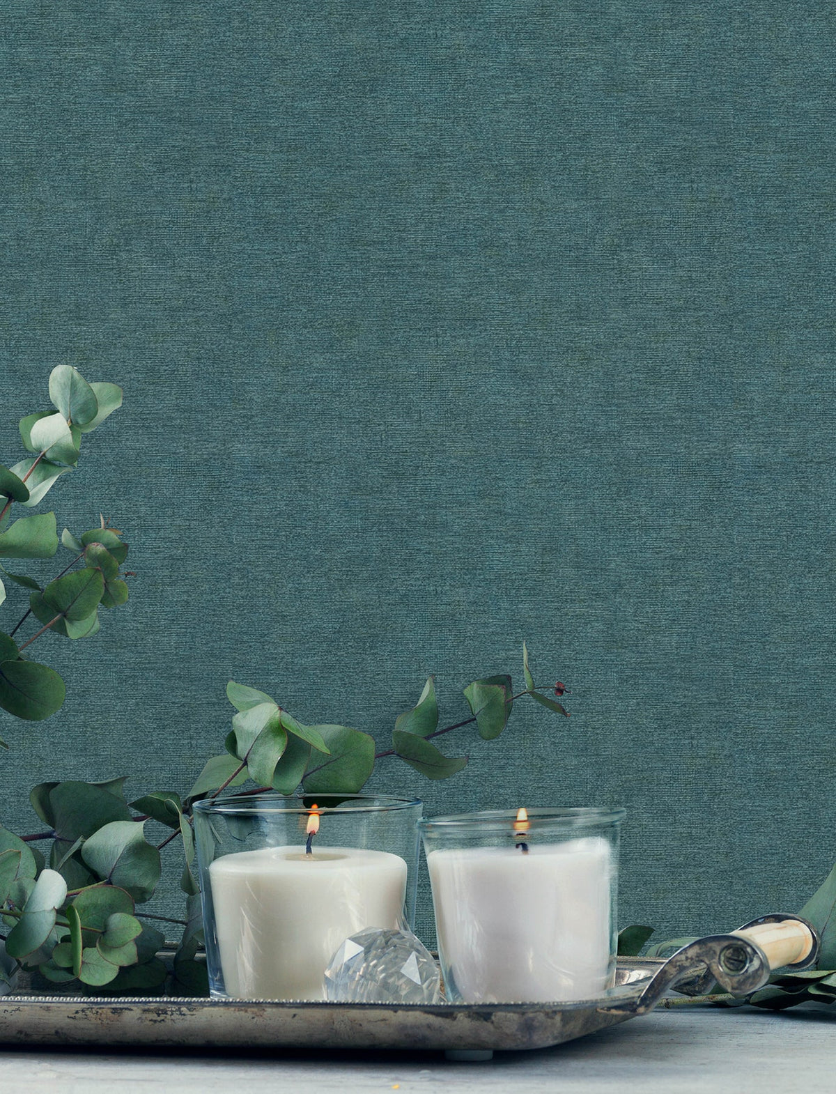 Two lit white candles in glass holders sit on a silver tray with eucalyptus branches and a crystal decoration, set against Chesapeake Tansy Distressed Wallpaper by York Wallcoverings.