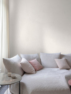 The serene space features Chesapeake Hollis Textured Diamond Wallpaper by York Wallcoverings, complementing a cozy, light-toned sofa with textured upholstery. Soft pink and gray throw pillows add warmth, while a small round side table with two white cups rests nearby. Sheer curtains filter gentle natural light, enhancing the calm and inviting ambiance.