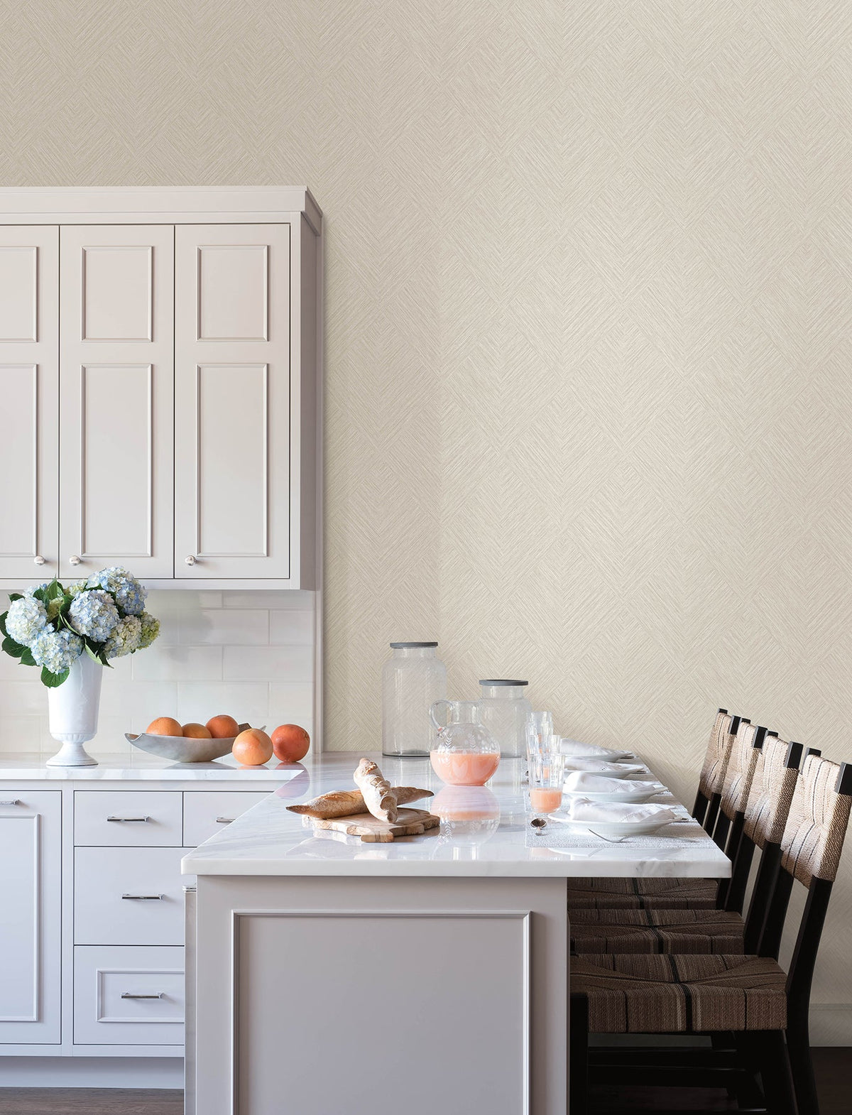 Modern kitchen with white cabinets, marble countertop, four wooden chairs, a vase of flowers, a bowl of oranges, glass containers, croissants, and a pitcher and glass of pink juice on the island. The space features Chesapeake Hollis Textured Diamond Wallpaper by York Wallcoverings as the neutral patterned background.