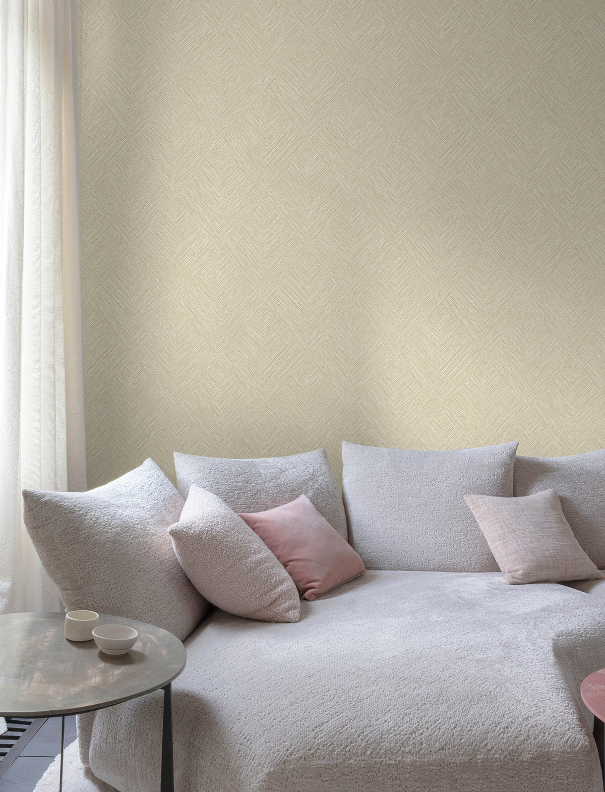 A modern living room features Chesapeake Hollis Textured Diamond Wallpaper by York Wallcoverings, adding a subtle pattern to the walls, along with a light gray textured sofa with pink and gray throw pillows, a round side table with two cups, and sheer white curtains letting in natural light.