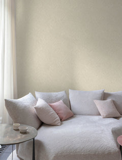 A cozy, light gray sectional sofa with textured fabric sits by a window adorned with sheer white curtains, set against Chesapeake Hollis Textured Diamond Wallpaper by York Wallcoverings. Pink and gray accent pillows are arranged on the sofa, next to a round side table holding two small white cups.