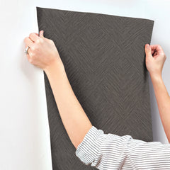 A person with rolled-up sleeves applies Chesapeake Hollis Textured Diamond Wallpaper by York Wallcoverings—a dark, textured wallpaper with a geometric diamond pattern—to a white wall, smoothing it with both hands.