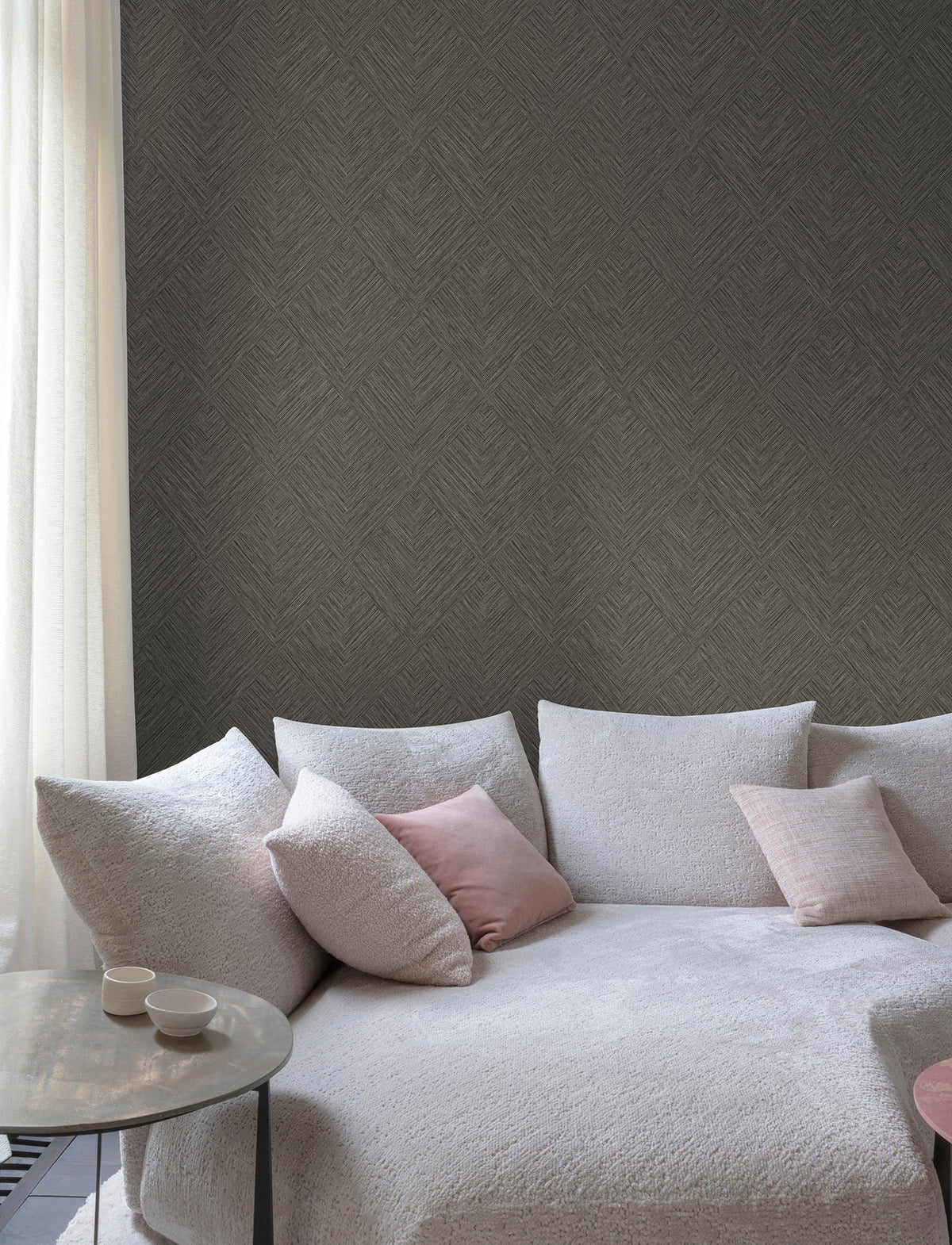 A cozy light gray sofa with textured fabric, pastel pink and cream cushions, a small round side table holding two cups, and a sheer white curtain sits beside a wall adorned with Chesapeake Hollis Textured Diamond Wallpaper by York Wallcoverings.