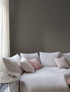 A cozy light gray sofa with textured fabric, pastel pink and cream cushions, a small round side table holding two cups, and a sheer white curtain sits beside a wall adorned with Chesapeake Hollis Textured Diamond Wallpaper by York Wallcoverings.