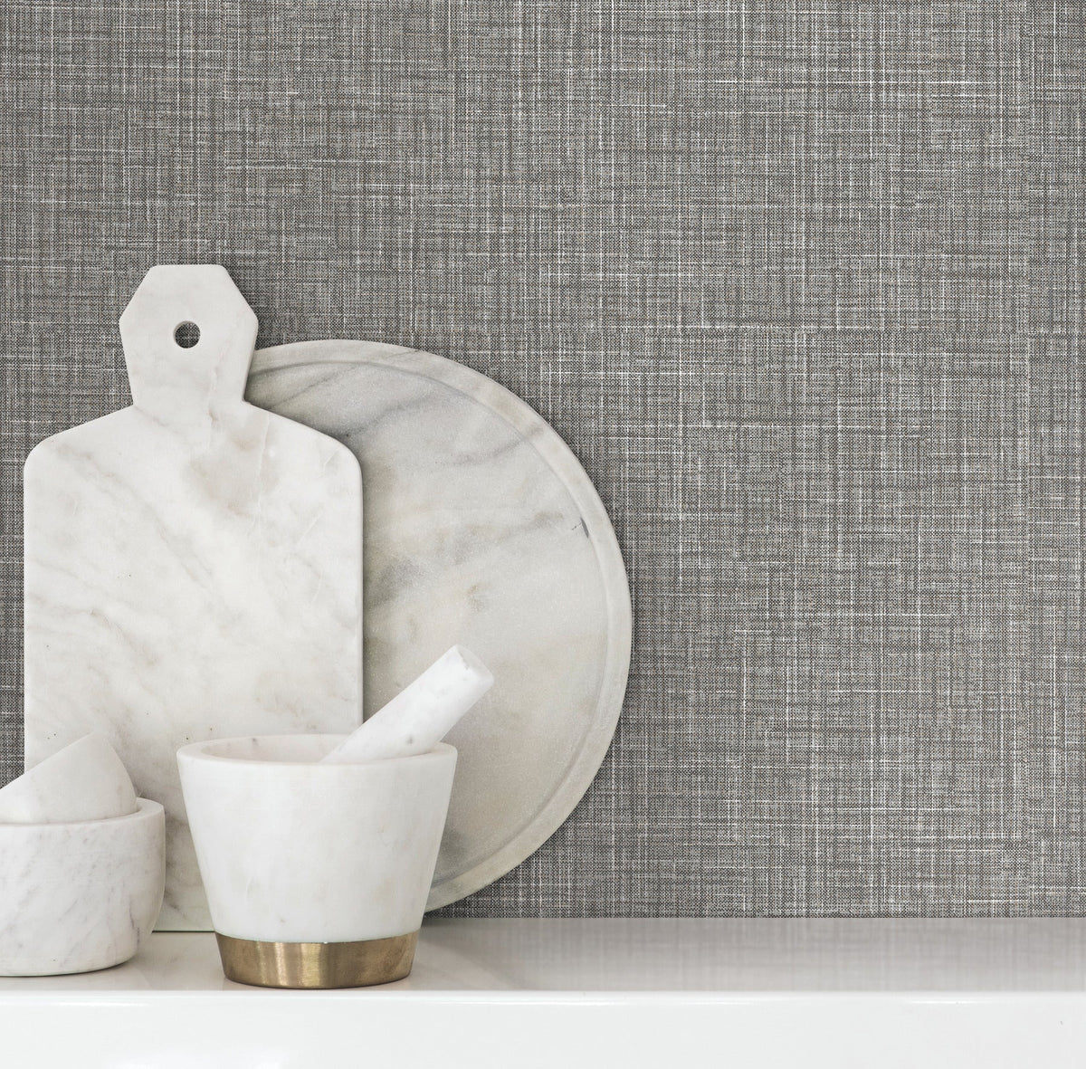 Beau Faux Fabric Wallpaper by York Wallcoverings creates a textured gray backdrop behind a white surface adorned with a marble mortar and pestle, round tray, and cutting board.