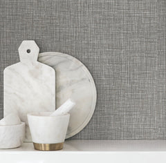 Beau Faux Fabric Wallpaper by York Wallcoverings creates a textured gray backdrop behind a white surface adorned with a marble mortar and pestle, round tray, and cutting board.