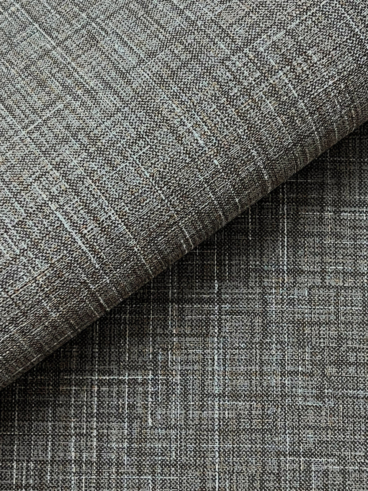 Close-up of Beau Faux Fabric Wallpaper by York Wallcoverings, showcasing a textured gray surface with a woven, crisscross pattern. One section is folded over, revealing a subtle variation in shading and texture.